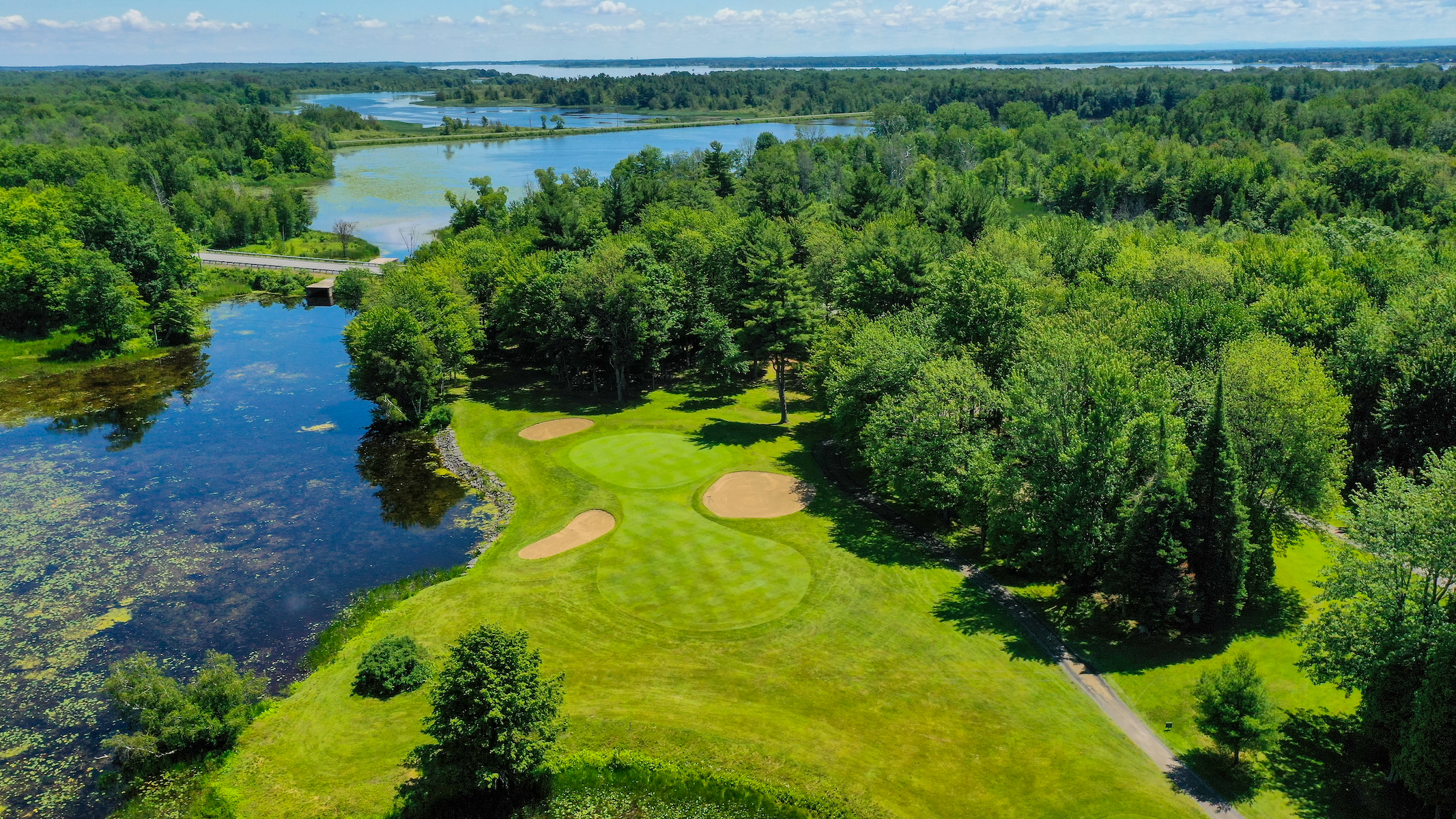 Aerial view of a lush Upper Canada Golf Course with multiple greens and sand traps, nestled beside a scenic lake and surrounded by dense forests under a clear blue sky.